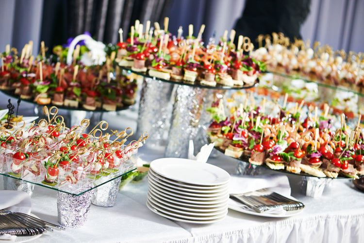Elegantly arranged buffet for corporate catering featuring a variety of gourmet dishes, canapés, and desserts, with a professional server in the background preparing plates for guests.